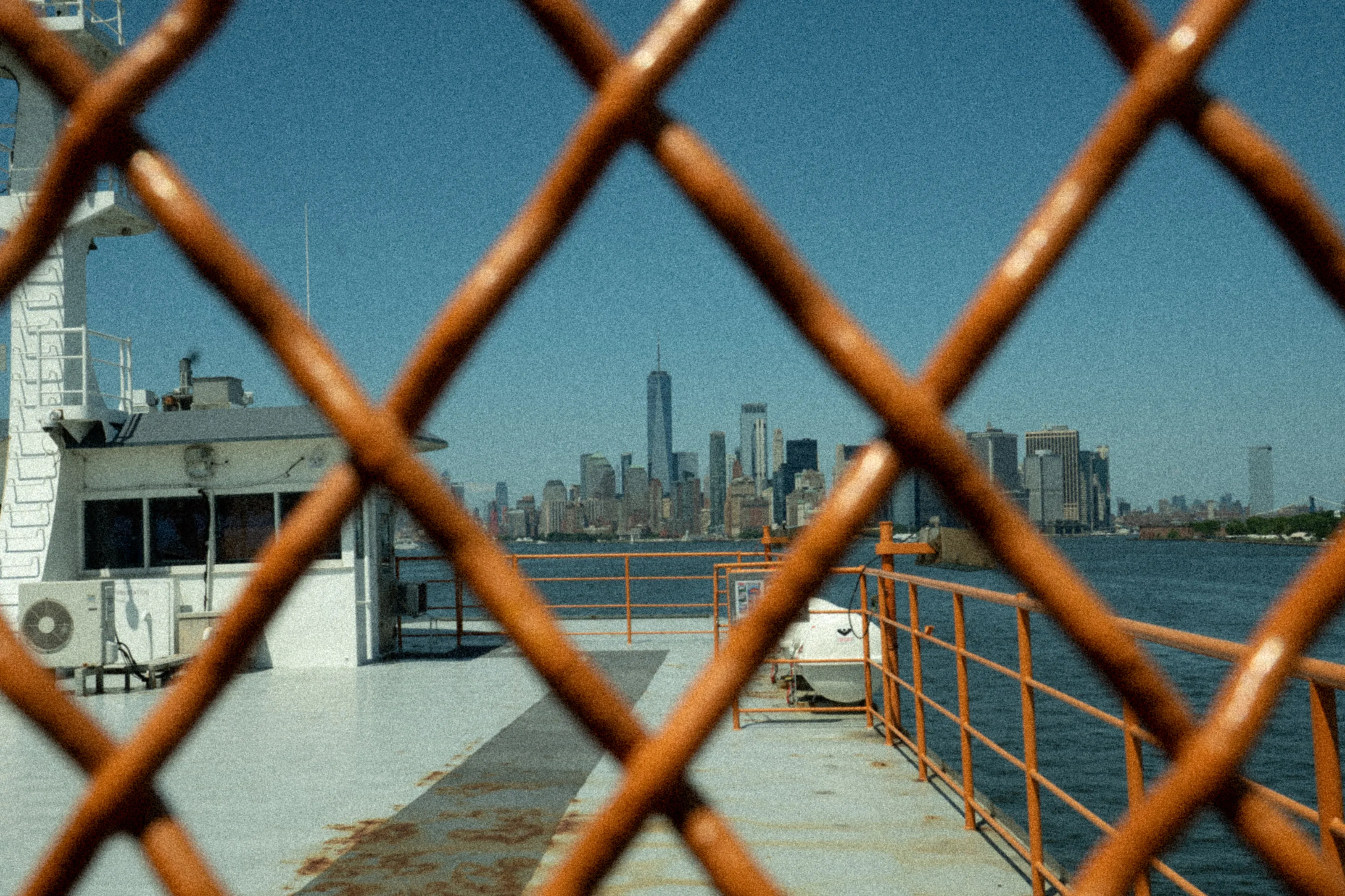Lower Manhattan seen through the orange diamond mesh of a ferry railing.