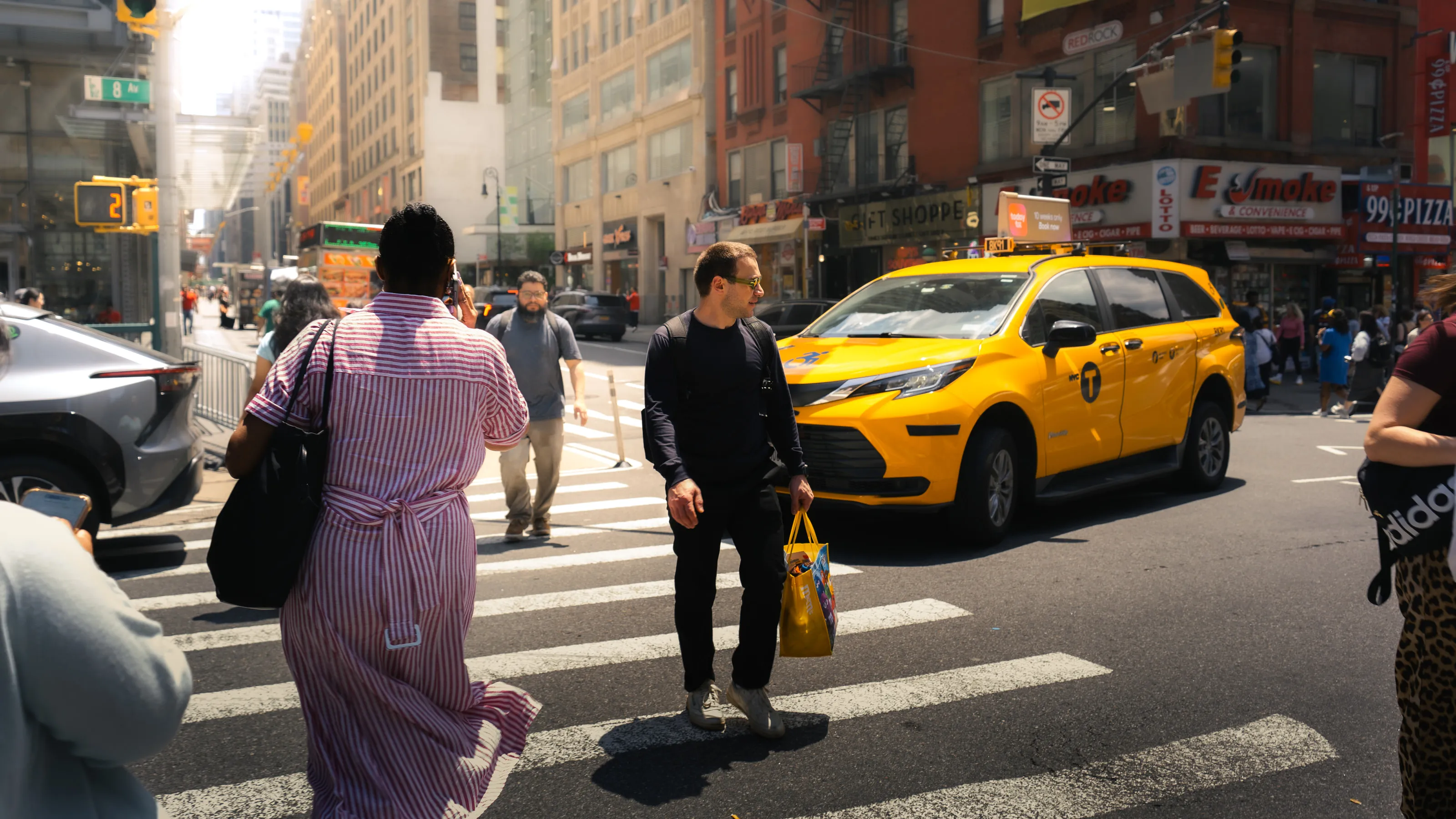 People cross a Midtown street in bright sun as a yellow cab waits at the light.