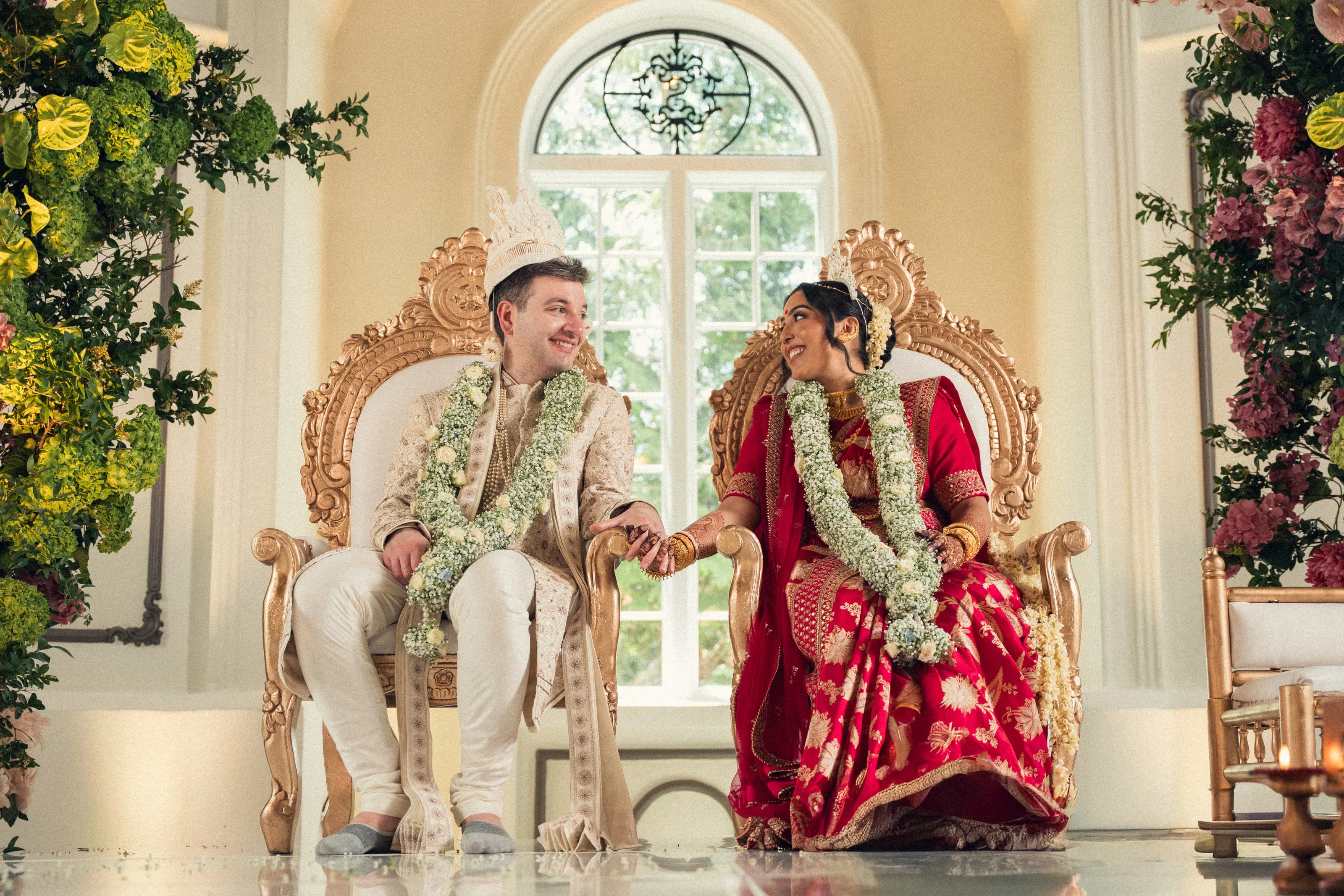 The couple sit on gilded chairs in garlands, holding hands beneath an arched window.
