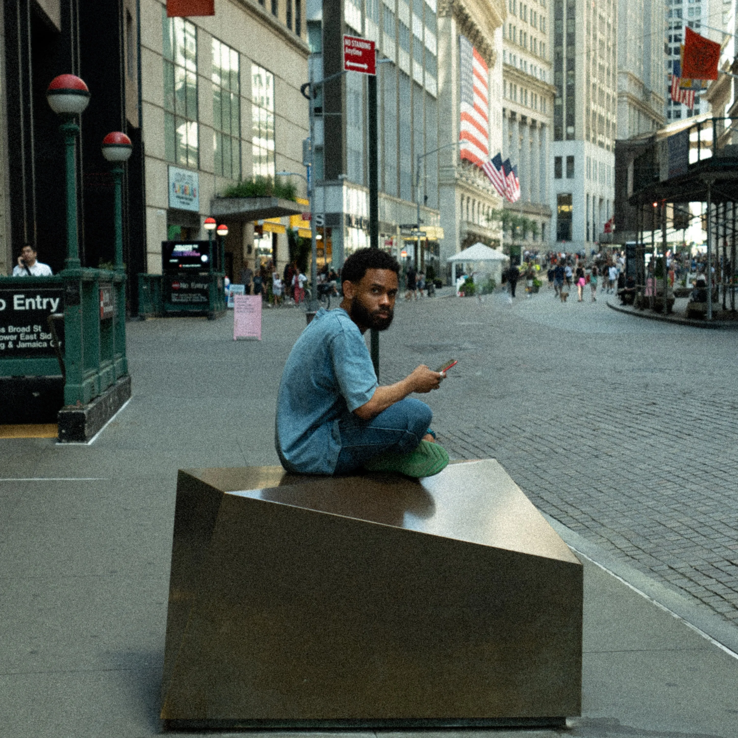 A man sits on a stone block looking at his phone, between office towers and flags.
