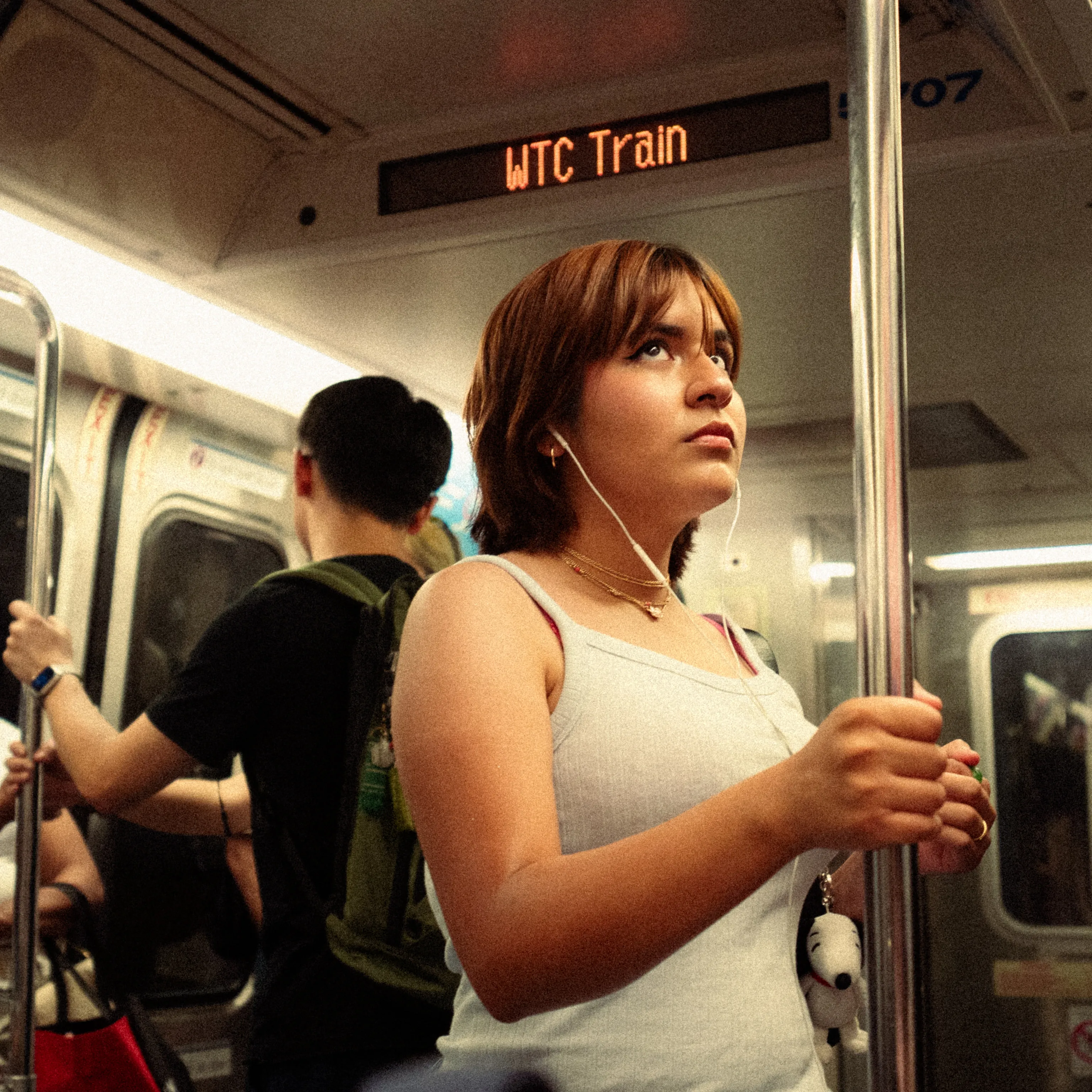 A young woman holds a pole on a train marked WTC, earphones in, looking up.