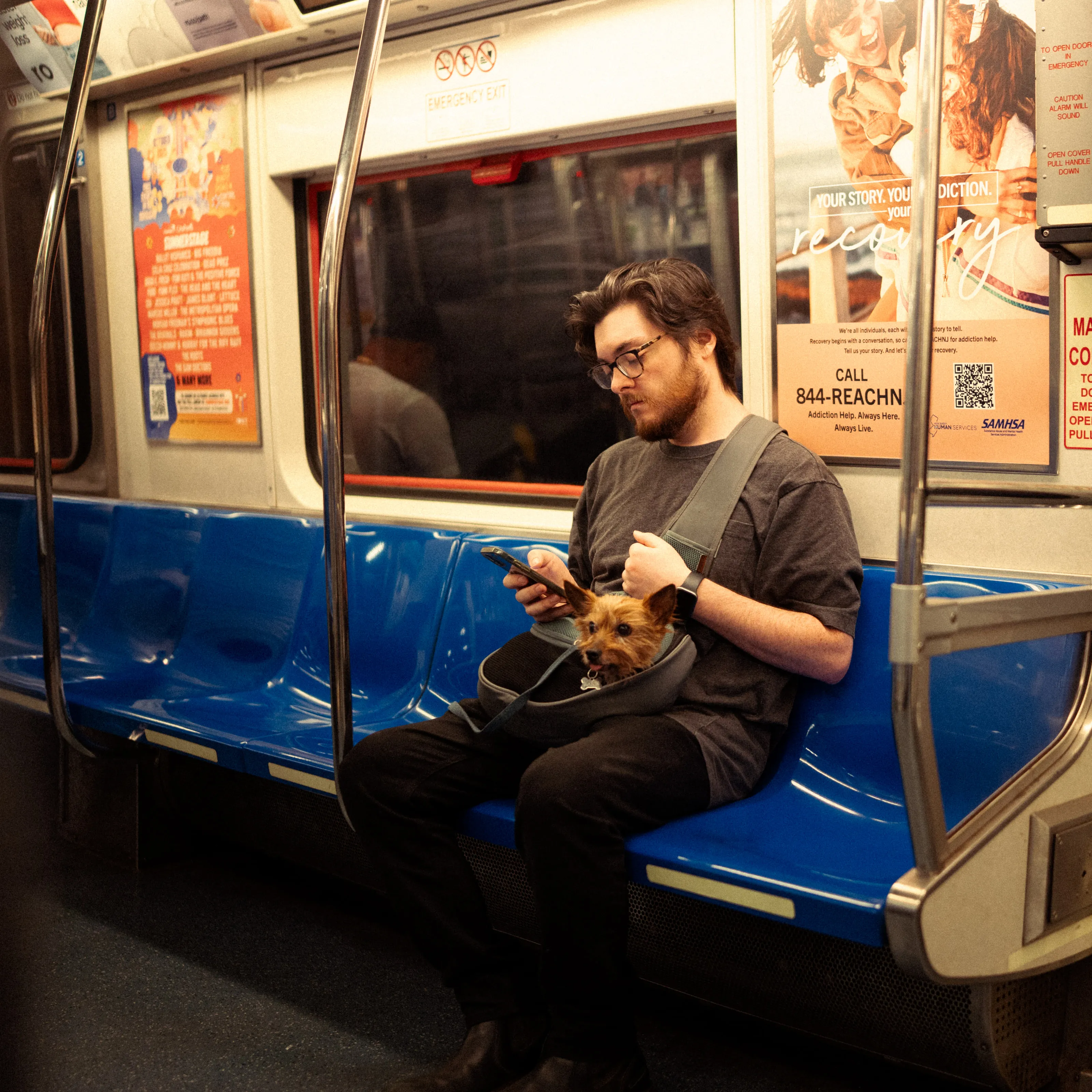 A man on a subway bench holds a small dog in a carrier on his lap.