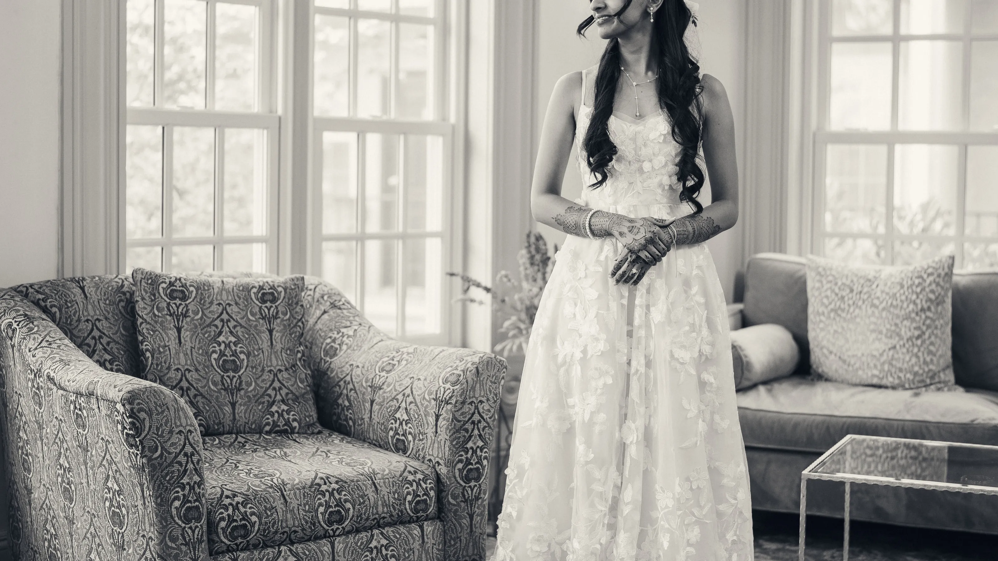 Black-and-white photograph of the bride in a lace gown in a sunlit sitting room.
