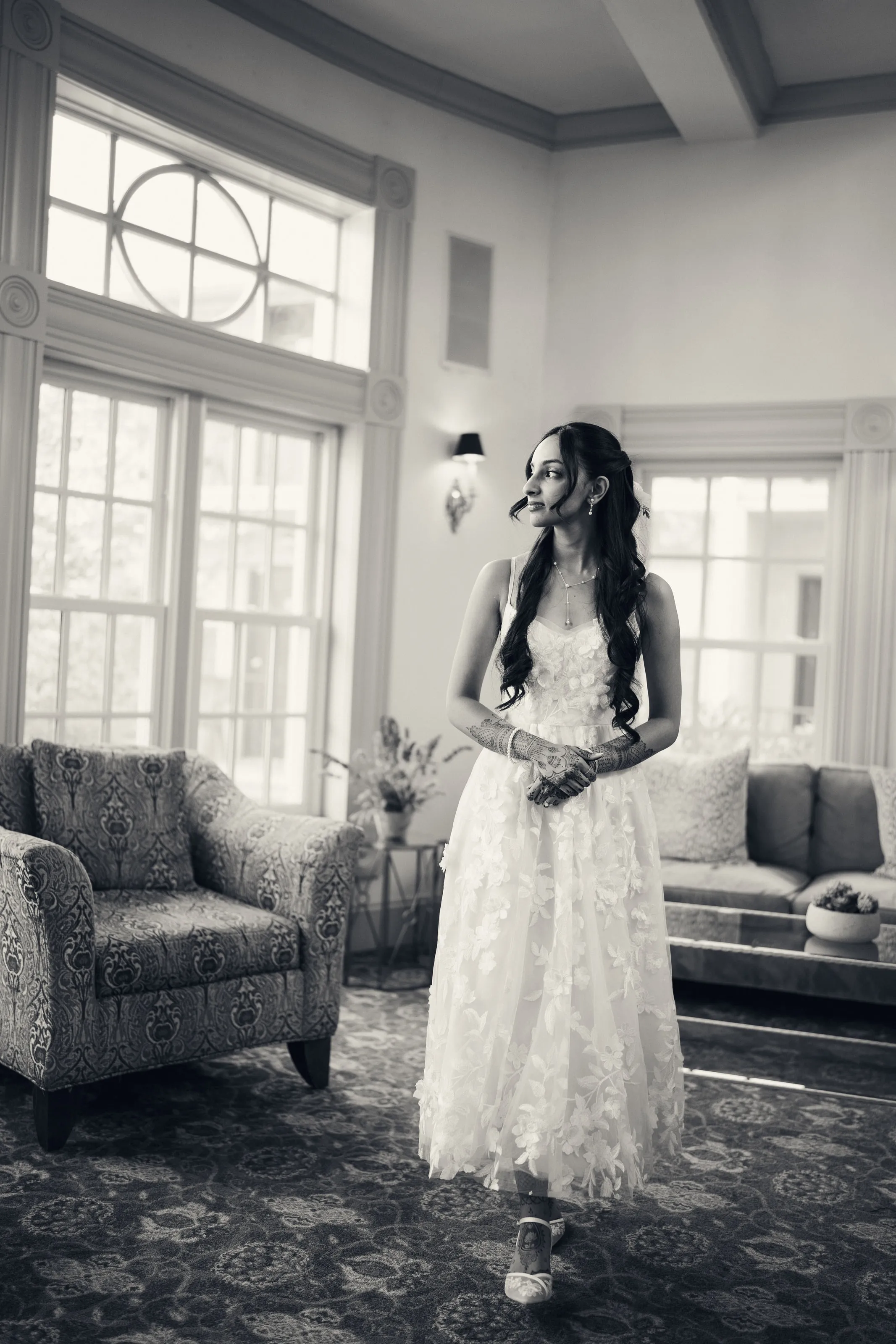 The bride walks through a sitting room with tall windows, in a lace gown.