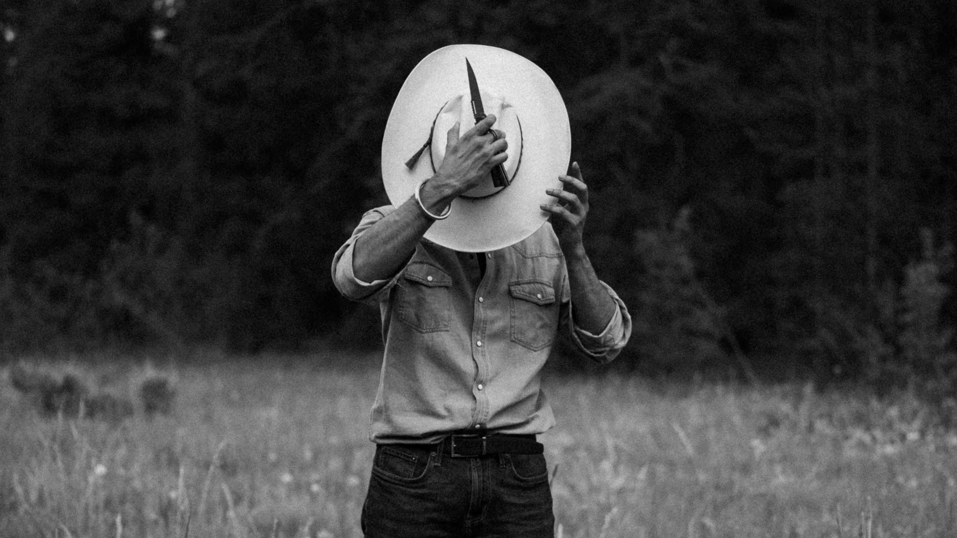 Black-and-white portrait of a man holding a cowboy hat over his face in a meadow.