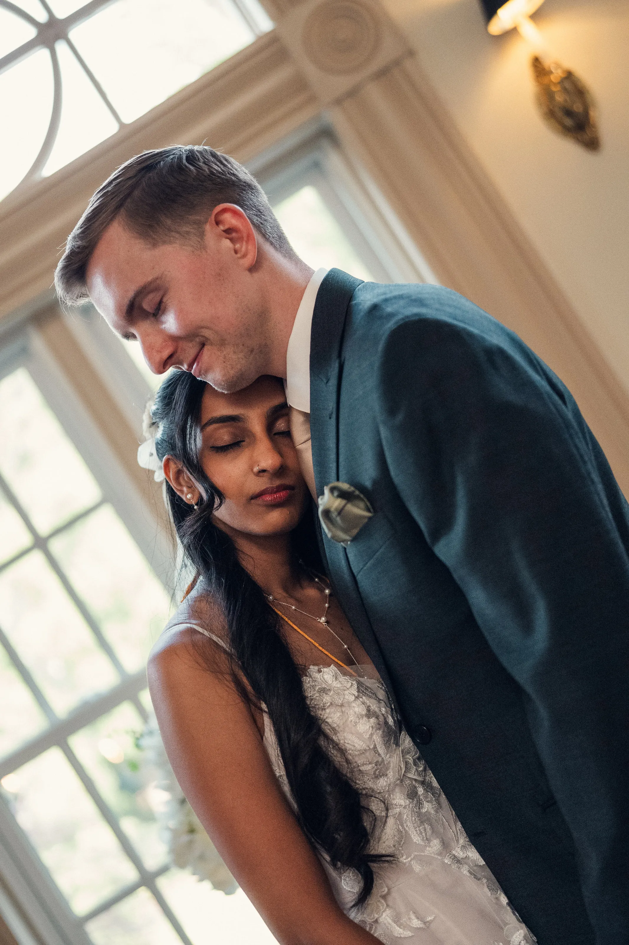 The groom holds the bride close, her eyes closed, in front of a window.