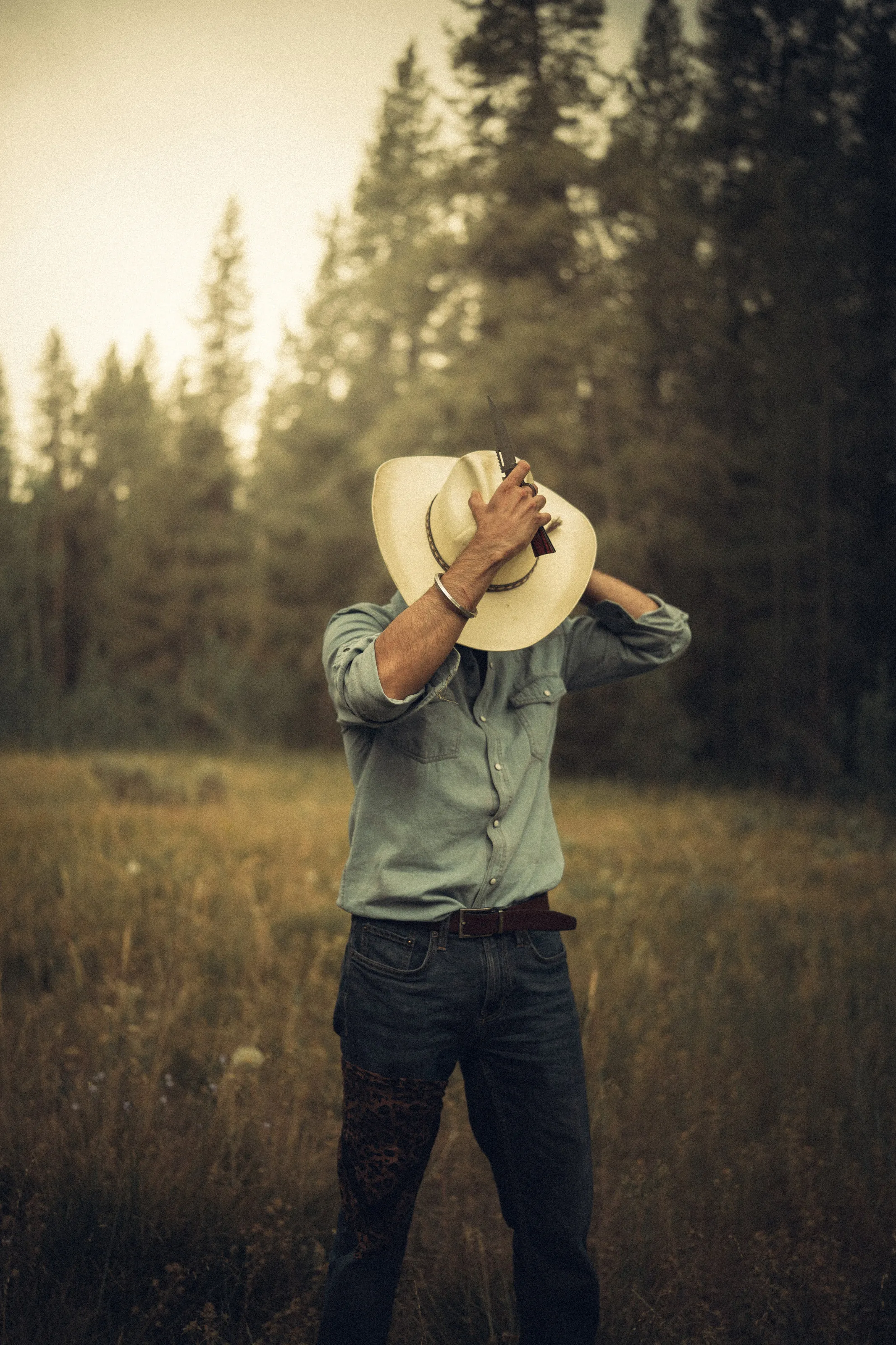 A man tips a cowboy hat over his face in a forest meadow.