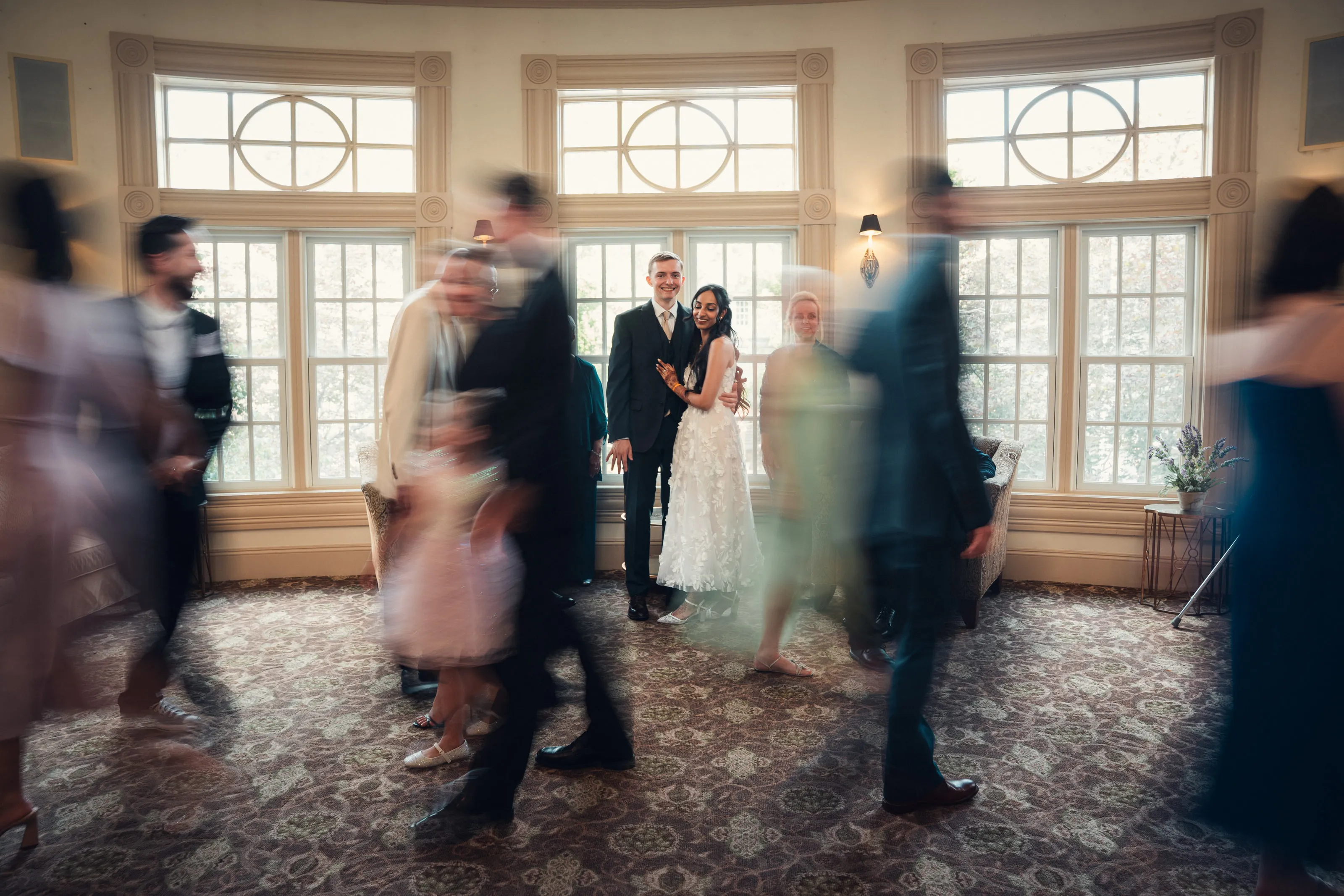 A long exposure of guests dancing around the couple in a room with tall windows.