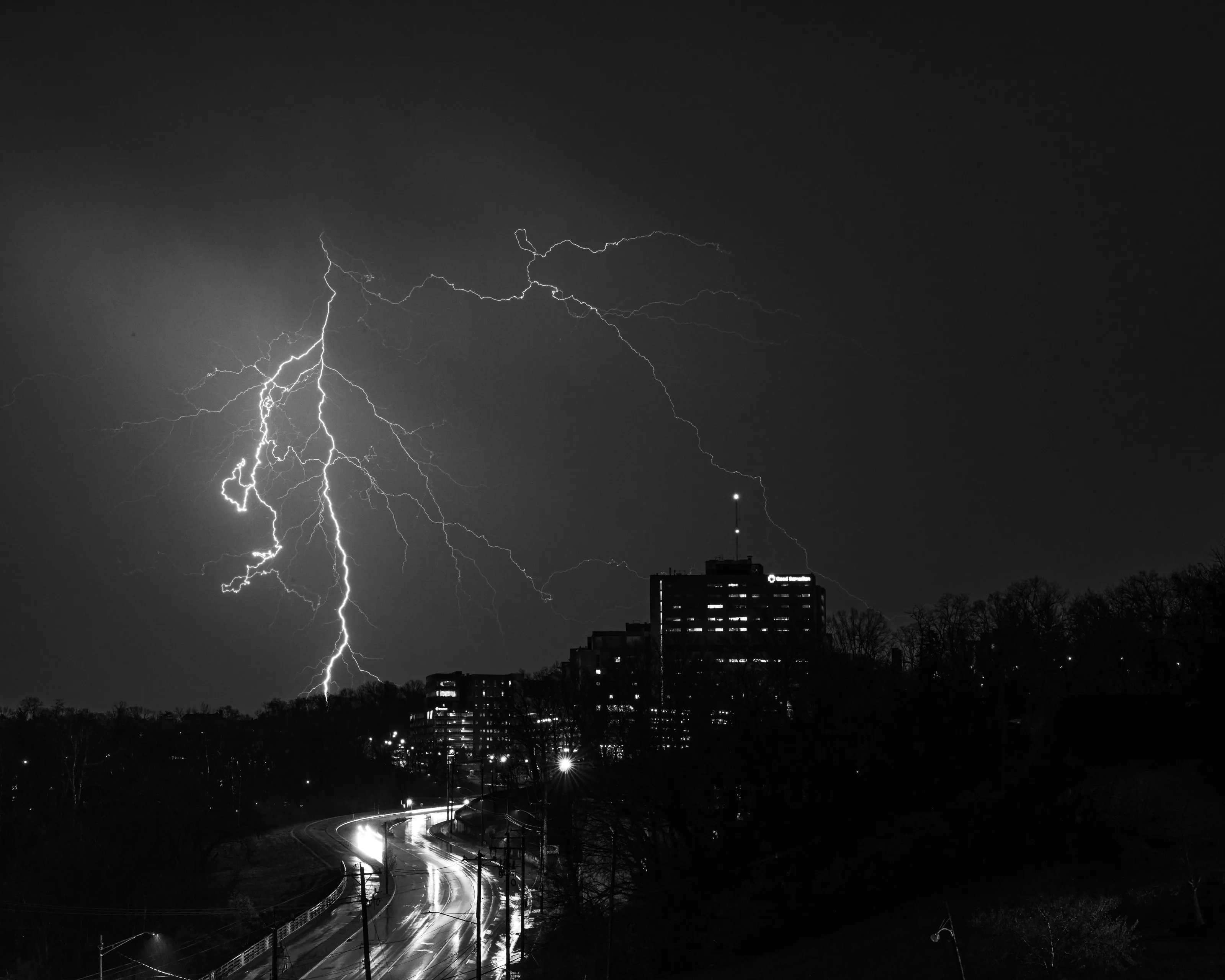 Black-and-white photograph of lightning forking over city towers at night, with light trails on a road below.