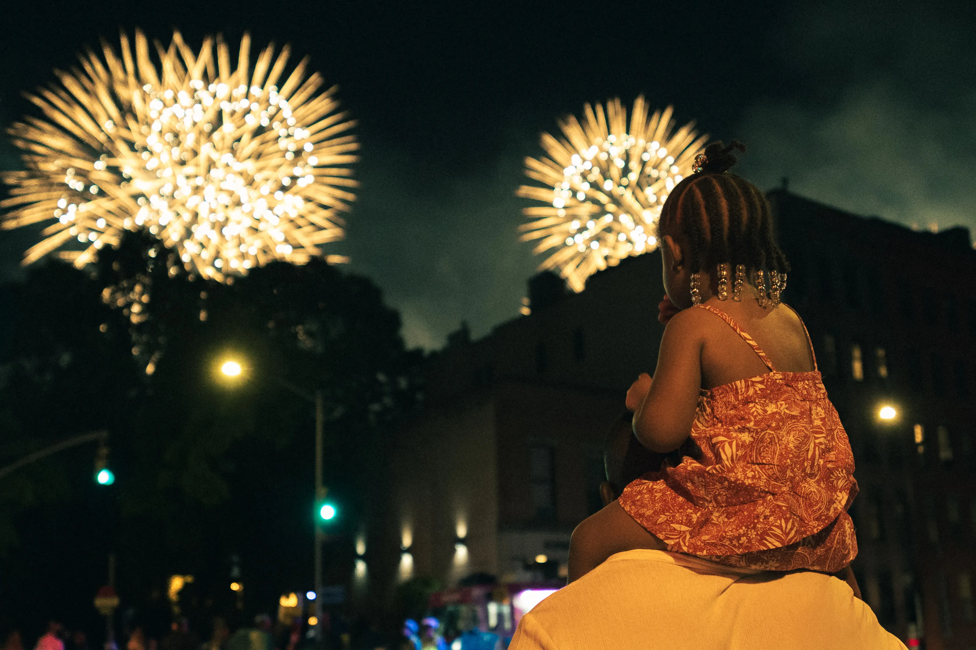 A child on someone's shoulders watches fireworks burst over the street.