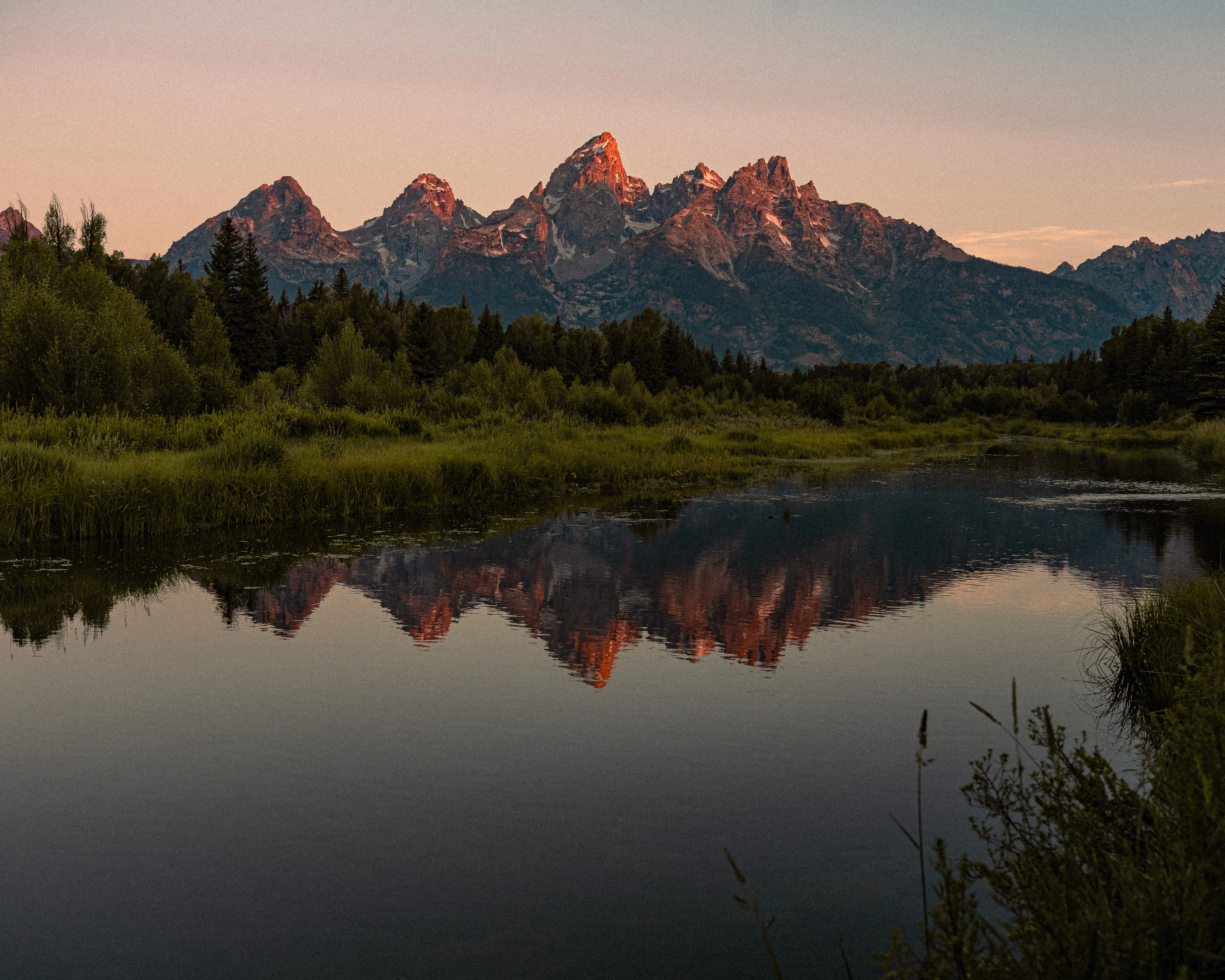 The Teton peaks glow pink at sunrise, mirrored in a still river.