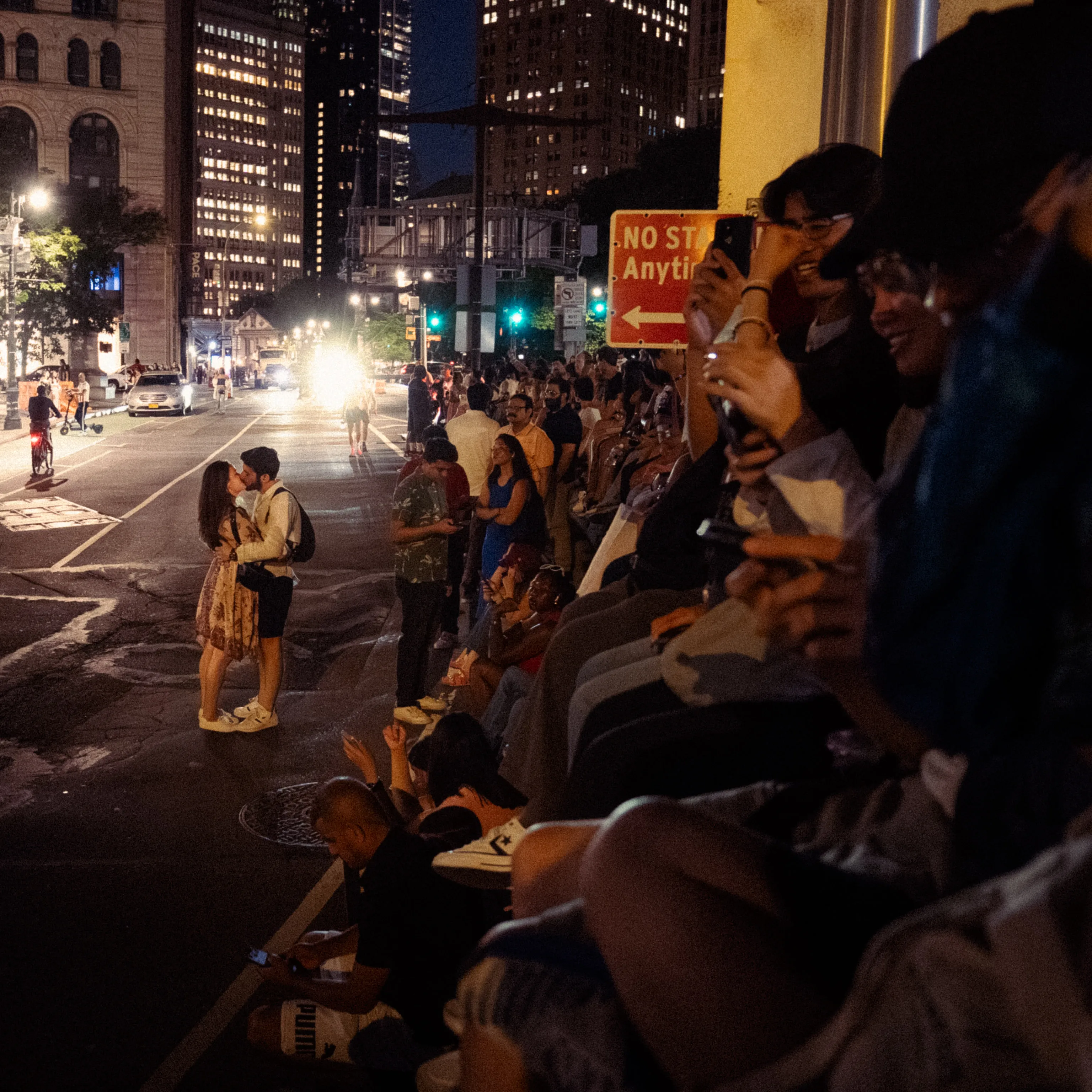 A couple kisses in the street at night beside a crowd sitting on the curb.