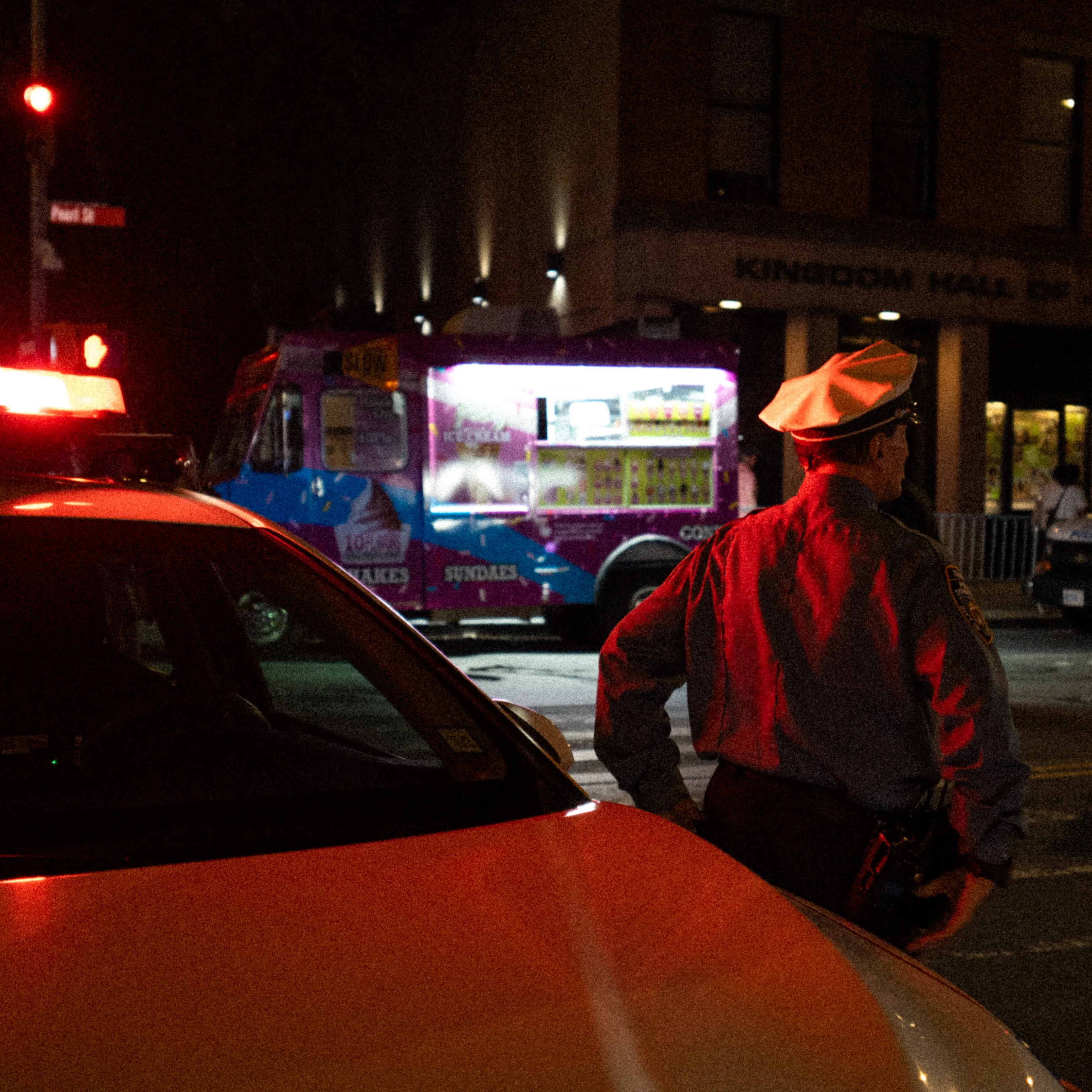 A police officer stands by a red car as an ice cream truck glows behind him at night.