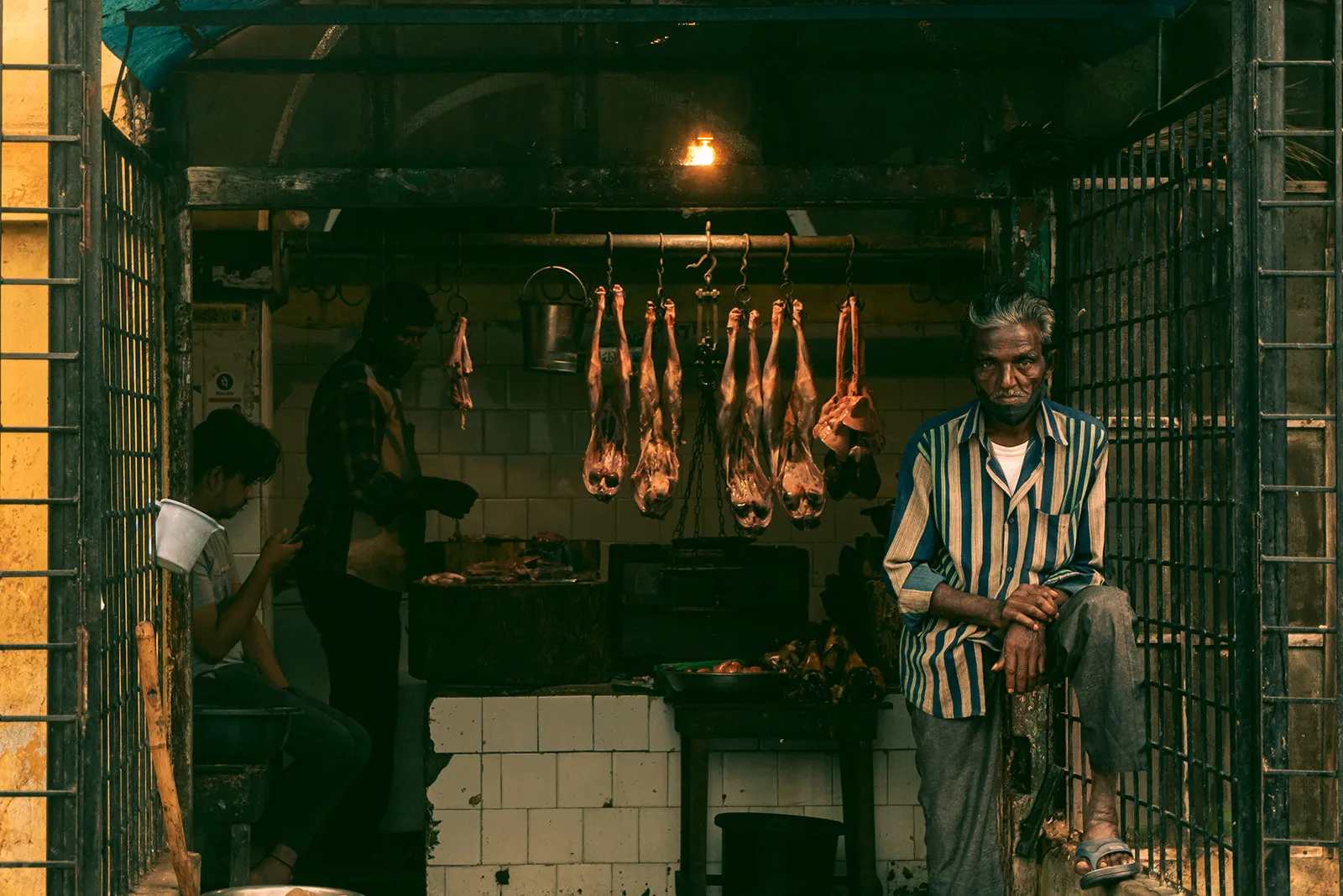 A butcher sits in a green-tiled stall with meat hanging behind him.
