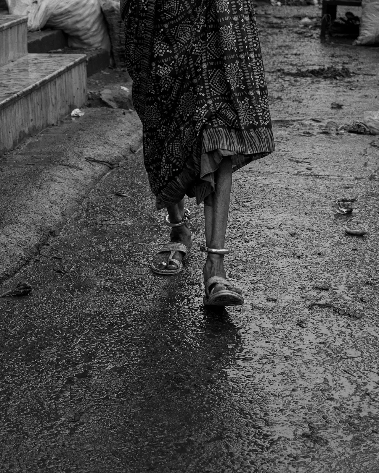 Black-and-white view of a woman's feet in sandals walking on wet pavement.